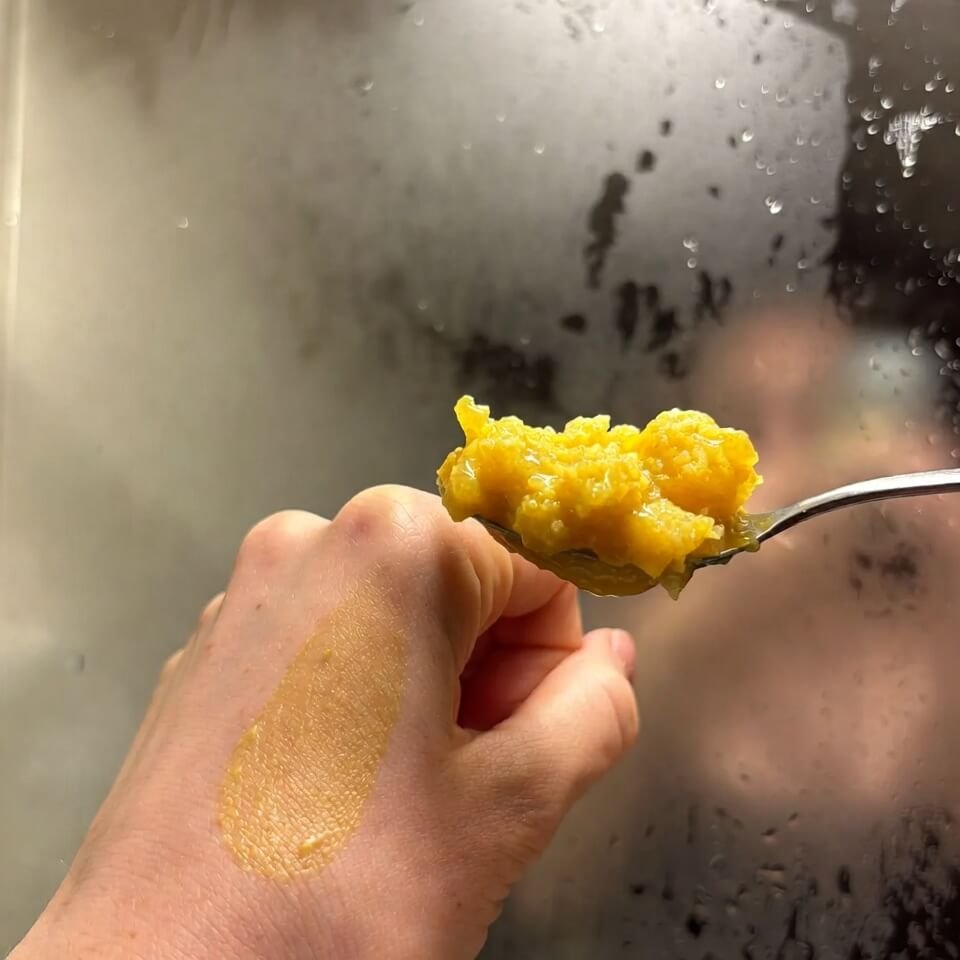 Hand holding messy yellow honey-aloe-turmeric mask spoon, foggy mirror background.